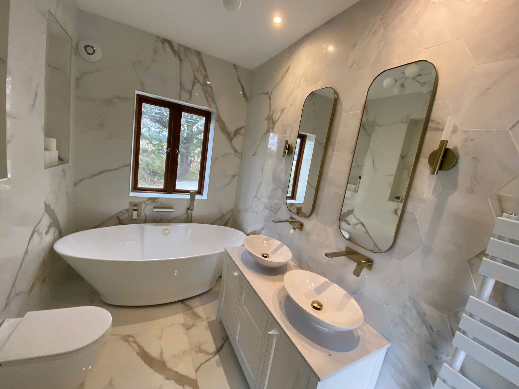 Calacatta marble ensuite in Stevenage with a freestanding oval bath beneath the window, twin vessel basins, brushed gold taps and slim gold framed mirrors